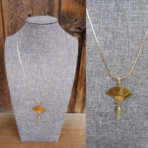 Puede incluir: Un collar dorado con un colgante en forma de abanico. El colgante tiene detalles intrincados y una pequeña borla colgando. El collar se exhibe en un maniquí gris sobre un fondo de madera.