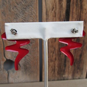 May include: A pair of red metal earrings with a zig-zag design. The earrings are on a white display stand.