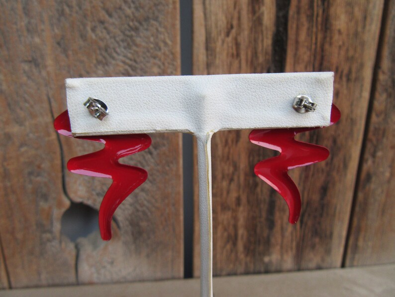 May include: A pair of red metal earrings with a zig-zag design. The earrings are on a white display stand.