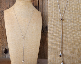 Peut inclure: Un collier lasso argenté composé d'une fine chaîne serpent qui passe à travers une petite perle en métal poli. Le collier se termine par deux breloques en forme de goutte d'eau de longueurs différentes, créant un design vertical élégant.