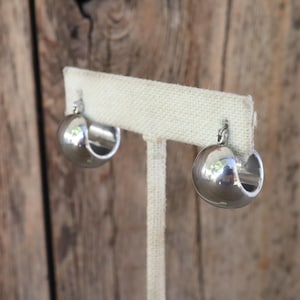 May include: A pair of polished silver hoop earrings. Each earring has a rounded, hollow design with a smooth, reflective surface. The earrings are displayed on a beige fabric stand against a blurred wooden background.
