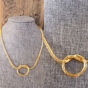 Könnte beinhalten: Eine goldfarbene Halskette mit Knotenmuster. Die Halskette wird auf einer grauen Schaufensterpuppe präsentiert. Die Kette besteht aus kleinen, rechteckigen Gliedern. Der Knoten ist ein kreisförmiges Design, ebenfalls aus denselben rechteckigen Gliedern.