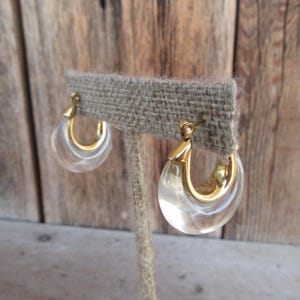 Puede incluir: Un par de pendientes de aro dorados con detalles de plástico transparente. Los pendientes se muestran en un soporte de arpillera.