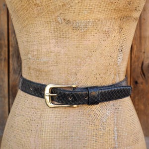 May include: A black belt with a gold-colored buckle is displayed on a mannequin. The belt has a textured, snake-skin pattern. The mannequin is set against a rustic wooden background.