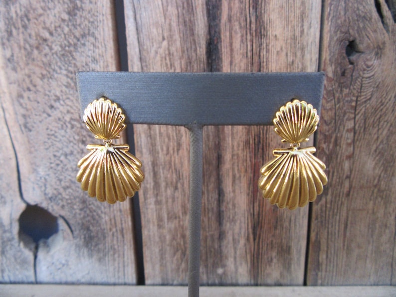 May include: A pair of gold-tone shell-shaped dangle earrings. The earrings are on a gray display stand.