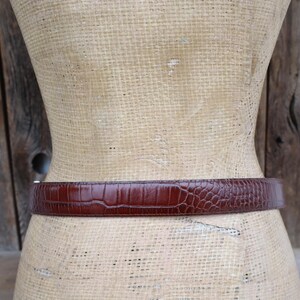 May include: A brown belt with a textured, crocodile-like pattern. The belt is wrapped around a mannequin. The background is a wooden wall.