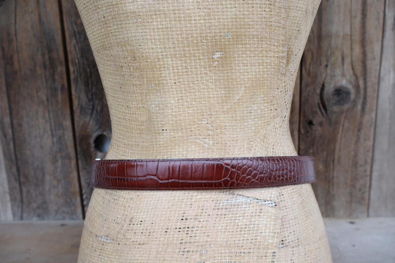 May include: A brown belt with a textured, crocodile-like pattern. The belt is wrapped around a mannequin. The background is a wooden wall.