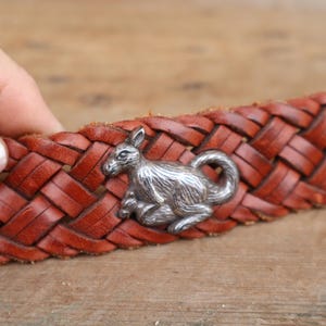 May include: A brown leather belt with a woven pattern. A silver kangaroo emblem is attached to the belt. The belt is held by a person's hand.