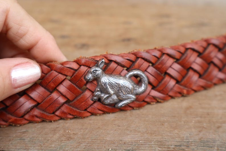 May include: A brown leather belt with a woven pattern. A silver kangaroo emblem is attached to the belt. The belt is held by a person's hand.