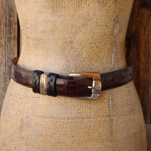 May include: A brown leather belt with a crocodile texture. The belt features a silver buckle with an ornate design and a braided leather accent. The belt is displayed on a mannequin.