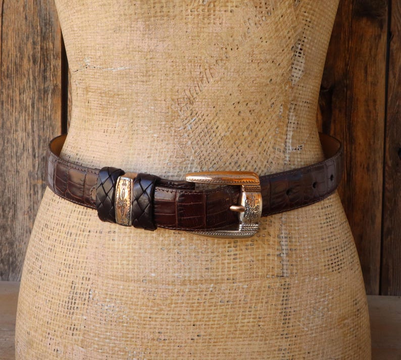 May include: A brown leather belt with a crocodile texture. The belt features a silver buckle with an ornate design and a braided leather accent. The belt is displayed on a mannequin.