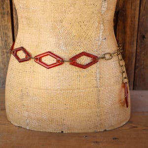 May include: A waist belt featuring a series of translucent amber-colored diamond-shaped plastic links connected by metallic gold-toned circular chain rings. The belt includes a dangling chain segment with a single diamond-shaped link at the end.