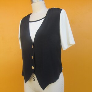 Peut inclure: Un gilet sans manches noir avec des boutons dor&eacute;s. Le gilet a une ourlet pointu et est port&eacute; sur un t-shirt blanc.