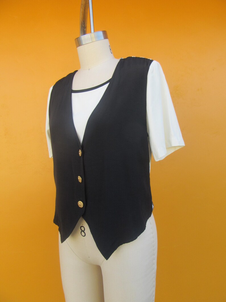 Peut inclure: Un gilet sans manches noir avec des boutons dor&eacute;s. Le gilet a une ourlet pointu et est port&eacute; sur un t-shirt blanc.