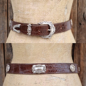 Op de afbeelding: Twee bruine leren riemen met zilverkleurige gespen en versieringen. De bovenste riem heeft een decoratieve gesp en bijpassende accenten. De onderste riem heeft een rechthoekige gesp en cirkelvormige accenten.