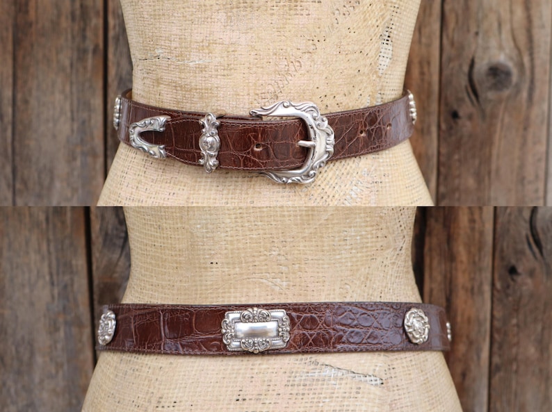 Op de afbeelding: Twee bruine leren riemen met zilverkleurige gespen en versieringen. De bovenste riem heeft een decoratieve gesp en bijpassende accenten. De onderste riem heeft een rechthoekige gesp en cirkelvormige accenten.