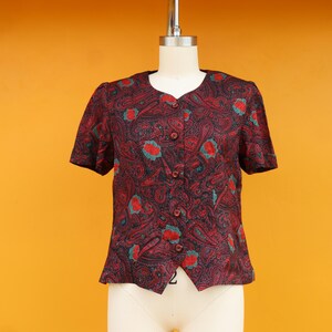 May include: A short-sleeved blouse with a paisley pattern in shades of burgundy, navy, and teal. The blouse has a button-up front and a rounded neckline. The garment is displayed on a mannequin against a yellow background.