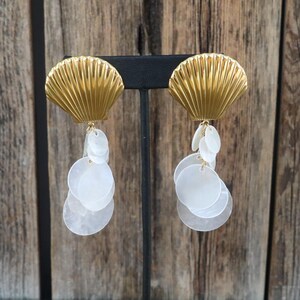 May include: Gold-toned seashell earrings with dangling white, circular accents. The earrings have a textured, ridged design on the shell portion and are displayed on a black stand against a wooden background. These earrings are a statement piece.