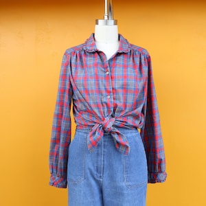 Op de afbeelding: Een rood, blauw en grijs geruit shirt met lange mouwen, een kraag met ruches en manchetten. Het shirt is in de taille geknoopt en gecombineerd met een jeansbroek met hoge taille. De outfit wordt op een mannequin gepresenteerd.