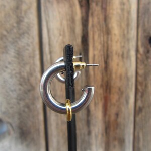May include: Silver hoop earrings with a gold accent. The earrings are on a black display stand.