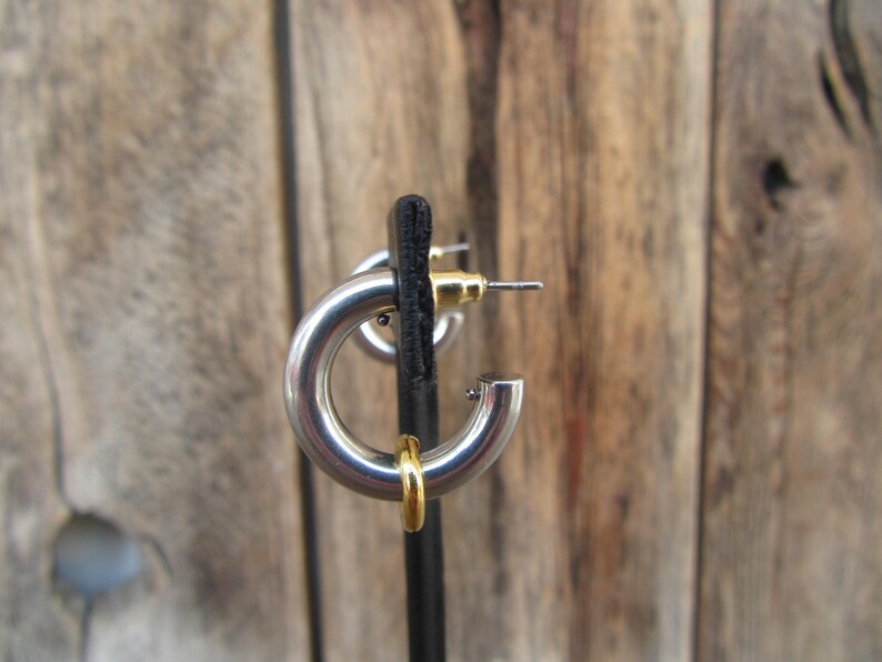 May include: Silver hoop earrings with a gold accent. The earrings are on a black display stand.