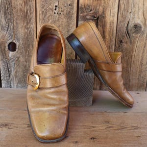 Puede incluir: Un par de zapatos vintage de cuero color canela con punta cuadrada y hebilla dorada. Los zapatos tienen un tacón bajo y se exhiben sobre una superficie de madera contra un fondo de madera. Los zapatos miden aproximadamente 28 cm de largo.