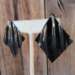 May include: Pair of black, diamond-shaped earrings with a glossy finish. Each earring features a textured, ridged design, creating a play of light and shadow. The earrings are displayed on a white stand against a wooden background.