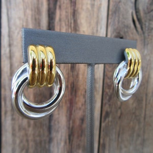 Op de afbeelding: Een paar zilver- en goudkleurige oorbellen. Elke oorbel heeft een dubbel lusontwerp met een zilverkleurige basis en een goudkleurig accent aan de bovenkant. De oorbellen worden getoond op een grijze standaard tegen een houten achtergrond.