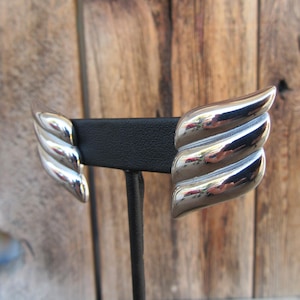 May include: A pair of silver earrings with a wing-like design. The earrings are on a black display stand.