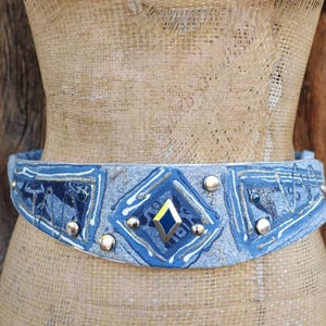 May include: A denim-style belt with a blue and silver design. The belt features a central diamond-shaped embellishment with a yellow and blue center, surrounded by silver studs. The belt is displayed on a mannequin.