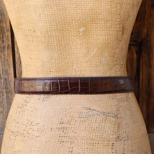 May include: A brown leather belt with a crocodile skin pattern is wrapped around a mannequin. The belt is a dark shade of brown and has a textured surface. The belt is a classic accessory.