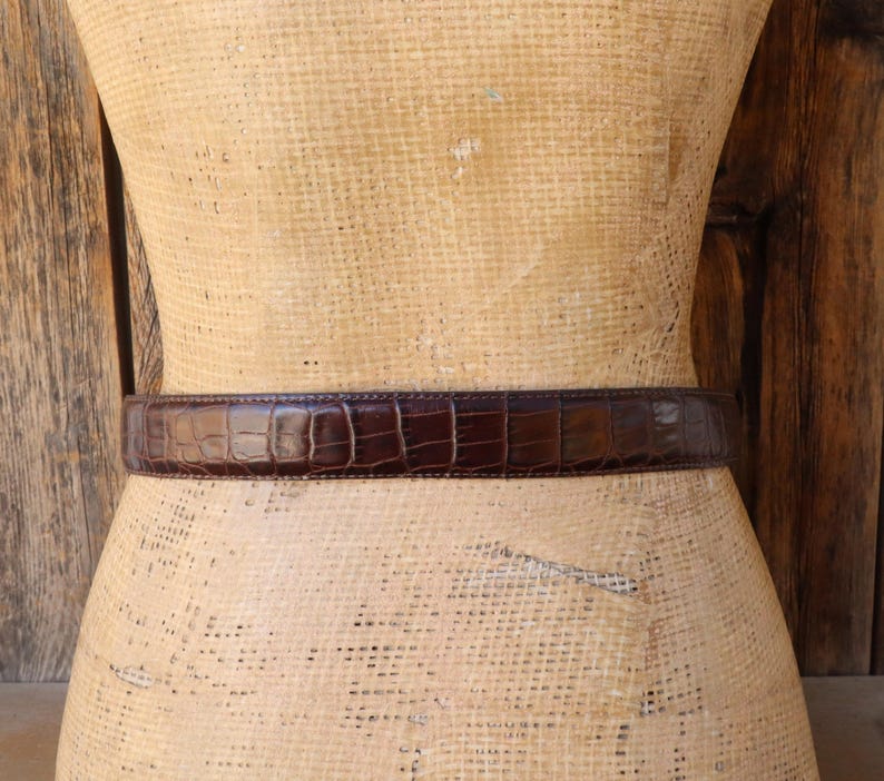 May include: A brown leather belt with a crocodile skin pattern is wrapped around a mannequin. The belt is a dark shade of brown and has a textured surface. The belt is a classic accessory.
