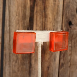 May include: A pair of orange square-shaped glass earrings. The earrings are on a white display stand.