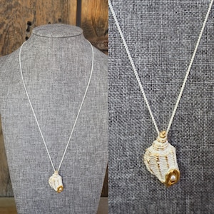 May include: A white and gold seashell pendant necklace. The pendant is a detailed seashell design with gold accents and a small pearl. The necklace has a delicate silver chain, displayed on a gray mannequin.