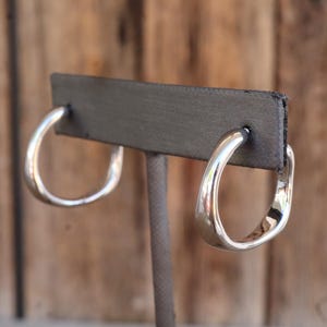 May include: A pair of silver-toned hoop earrings with a unique, slightly squared-off design. The earrings are displayed on a gray earring stand against a blurred wooden background. The hoops have a polished, reflective surface.