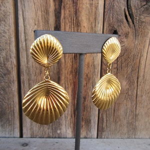 May include: A pair of gold tone dangle earrings with a shell design. The earrings are on a display stand.