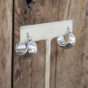 May include: A pair of shiny, silver-colored hoop earrings. The earrings are displayed on a white fabric earring holder against a blurred wooden background. The earrings have a rounded, half-circle design.