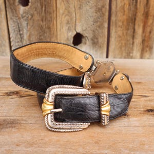 May include: A black leather belt with a silver and gold buckle. The belt has a textured pattern and a light brown interior. The buckle is rectangular with a gold center and silver trim. The belt is displayed on a wooden surface.