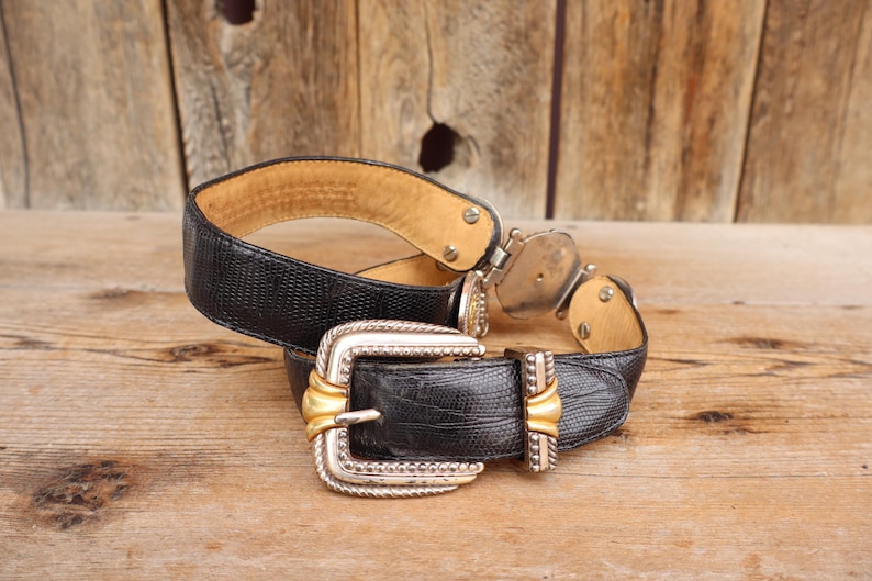May include: A black leather belt with a silver and gold buckle. The belt has a textured pattern and a light brown interior. The buckle is rectangular with a gold center and silver trim. The belt is displayed on a wooden surface.