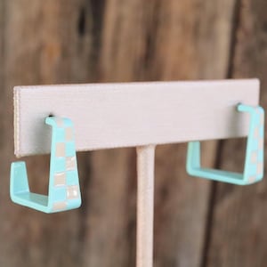 May include: A pair of square-shaped hoop earrings featuring a light blue and beige checkered pattern. The earrings have a smooth, glossy finish and a distinct geometric silhouette, displayed on a beige earring card for clear visibility.