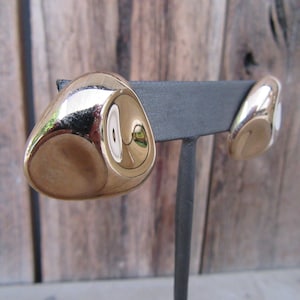 Può includere: Un paio di orecchini dorati a forma di goccia. Gli orecchini hanno una superficie liscia e riflettente e sono esposti su un espositore nero per orecchini. Lo sfondo è una superficie di legno invecchiato.