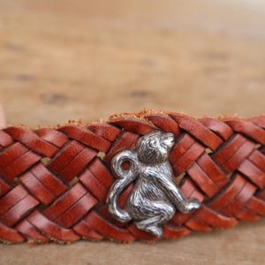 May include: A brown leather belt with a plaited design. A silver-coloured monkey charm is attached to the belt. The belt is made of woven leather and has a classic, rustic appearance.