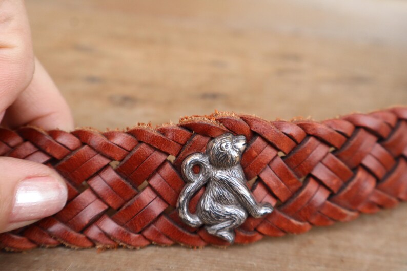 May include: A brown leather belt with a plaited design. A silver-coloured monkey charm is attached to the belt. The belt is made of woven leather and has a classic, rustic appearance.