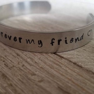Può includere: Un bracciale a polsino color argento con l'iscrizione "always forever my friend" in un carattere scritto a mano. Il bracciale aperto è appoggiato su una superficie di legno.