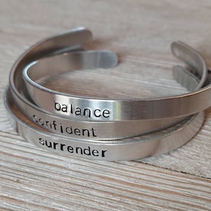 Puede incluir: Tres pulseras de puño de metal plateado apiladas sobre una superficie de madera. Las pulseras están grabadas con las palabras "balance", "confident" y "surrender" en una fuente sencilla. Las pulseras tienen un acabado de metal cepillado.