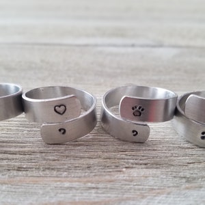 Puede incluir: Cuatro anillos de plata con diferentes diseños. Los anillos son abiertos y se pueden usar en cualquier dedo. Los diseños incluyen un corazón, una huella de pata, un punto y un signo de interrogación.
