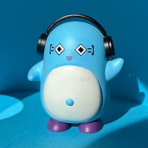 May include: A blue figurine of a cartoon character with a white belly, purple feet, and black headphones. The character has diamond-shaped eyes and a small purple mouth. The background is a solid blue.