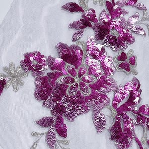 May include: A floral applique featuring magenta sequins and silver-colored embroidery on a white mesh backing. The design showcases a cluster of flowers and leaves, ideal for embellishing garments or accessories.