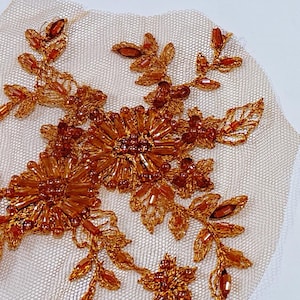 May include: Two decorative floral appliques. Each applique features a central flower design surrounded by leaves and smaller flowers, all in shades of amber and gold. The embellishments are attached to a beige mesh backing.