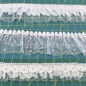 May include: Three different white lace trims are displayed against a green grid background. The top trim features scalloped edges and small pearls. The middle trim has a dotted pattern and small pearls. The bottom trim has floral and pearl details.